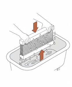 Plissee Nassreinigung Eine schematische Illustration zeigt die Handwäsche eines kompakten Wabenplissees in einer Badewanne. Zwei Hände halten die Schienen des Plissees fest, während es senkrecht in das Wasserbad eingetaucht wird. Ein brauner Pfeil, der nach unten zeigt, und ein brauner Pfeil, der nach oben zeigt, verdeutlichen das vorsichtige Schwenken des Stoffes im Wasser, um Verschmutzungen zu lösen.