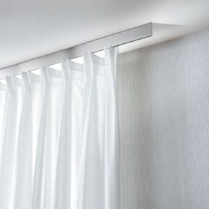 Puristisches Interstil Schienensystem „Spähre“ in Edelstahl, bündig an einer weißen Decke montiert. Die extrem flache, rechteckige Vorhangschiene trägt einen weißen, halbtransparenten Vorhang der in dekorativen Falten fällt. Das Bild betont eine helle, lichtdurchflutete Atmosphäre mit sanften Lichtreflexionen an der Decke.